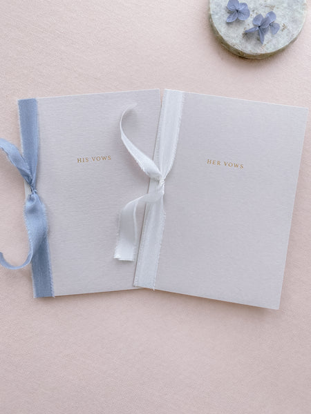 A set of two His and Her gold foil gray card stock vow books in typeface font with one tied in white colored silk ribbon and the other in blue colored silk ribbon