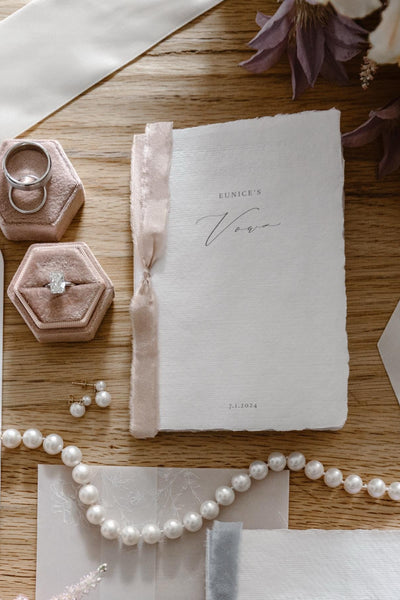wedding details flat lay showcasing white personalized wedding vow book with pink ribbon