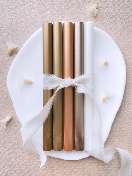 a set of 5 metallic earthy tones glue gun sealing wax sticks in gold, light gold, peachy gold, mocha, white pearl colors tied with soft white silk ribbon styled with dried flower petals