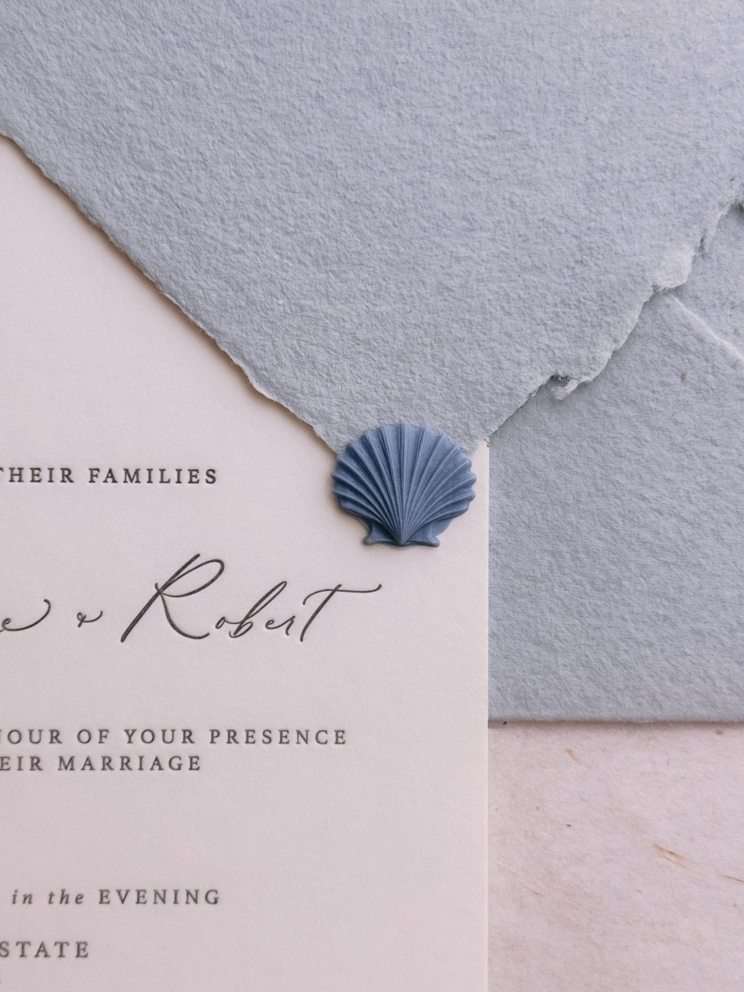Dusty blue 3D die-cut seashell wax seal on a white letterpress wedding invitation card and a light blue handmade paper envelope