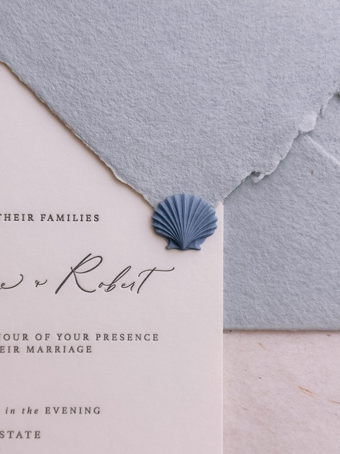 Dusty blue 3D die-cut seashell wax seal on a white letterpress wedding invitation card and a light blue handmade paper envelope