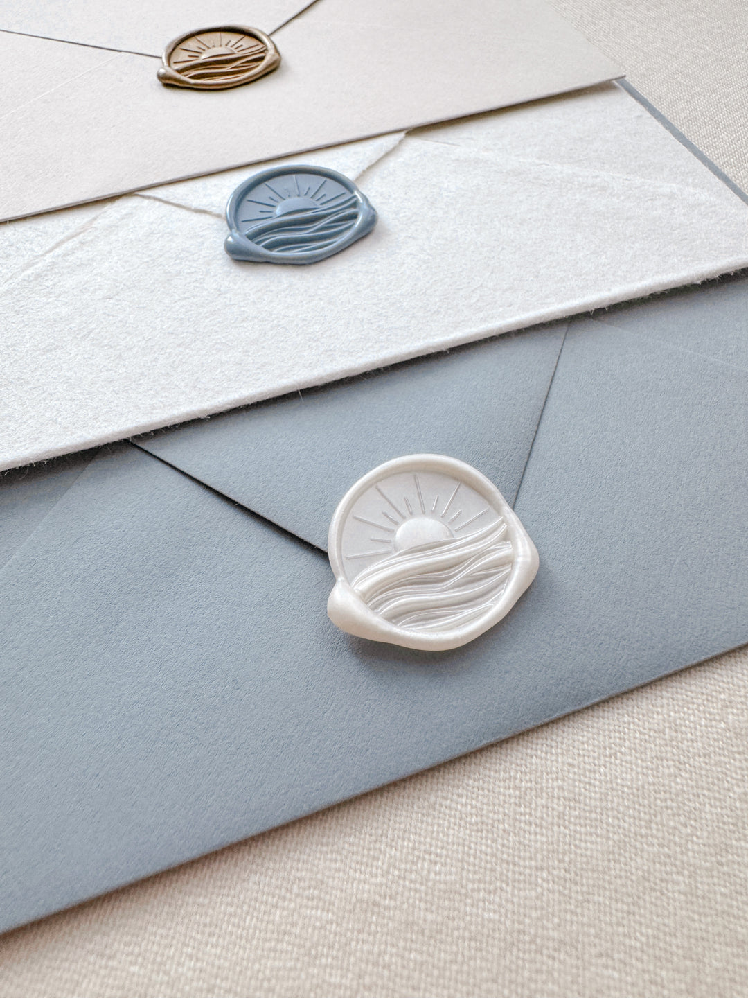 Pearl white, dusty blue, and gold 3D sunrise and ocean waves wax seals on beige, white deckle-edge, and dusty blue envelopes