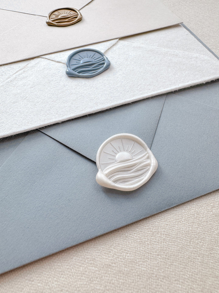Pearl white, dusty blue, and gold 3D sunrise and ocean waves wax seals on beige, white deckle-edge, and dusty blue envelopes