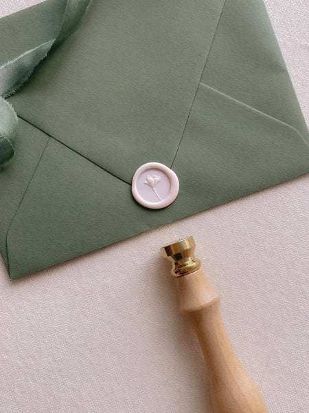 Light pink mini flower wax seal on an olive green envelope styled with a strand of green silk ribbon and wax seal stamp
