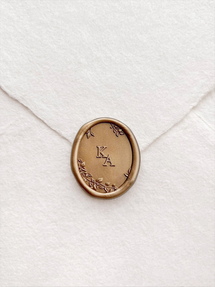 Golden bronze custom monogram wax seal featuring two initials framed by 3D engraved leaves and branches on a white handmade paper envelope