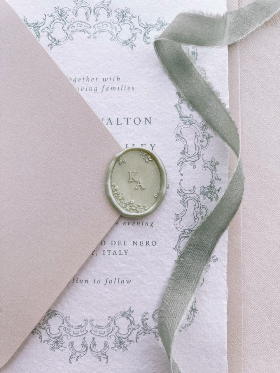 Light metallic sage green personalized initials oval wax seal on a beige envelope over a wedding invitation card