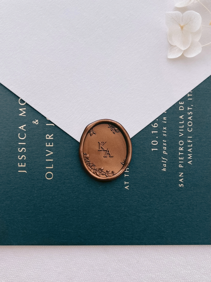 Bronze oval botanical monogram wax seal with custom initials on a beige envelope over a dark green wedding invitation card