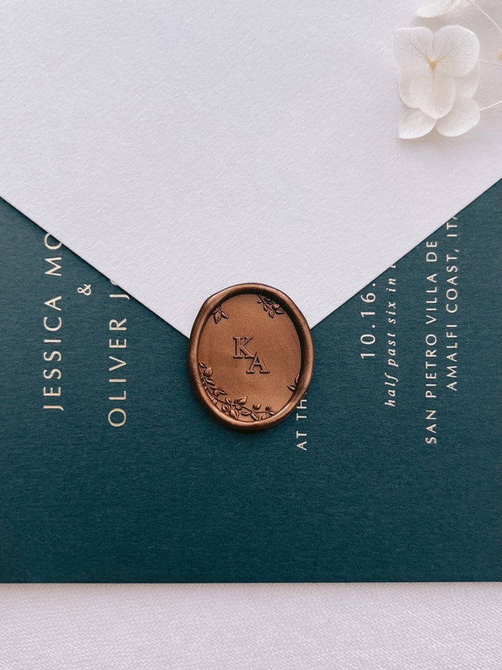 Bronze custom monogram oval wax seal with personalized initials and 3D engraved botanical elements on a beige envelope over a dark green wedding invitation card