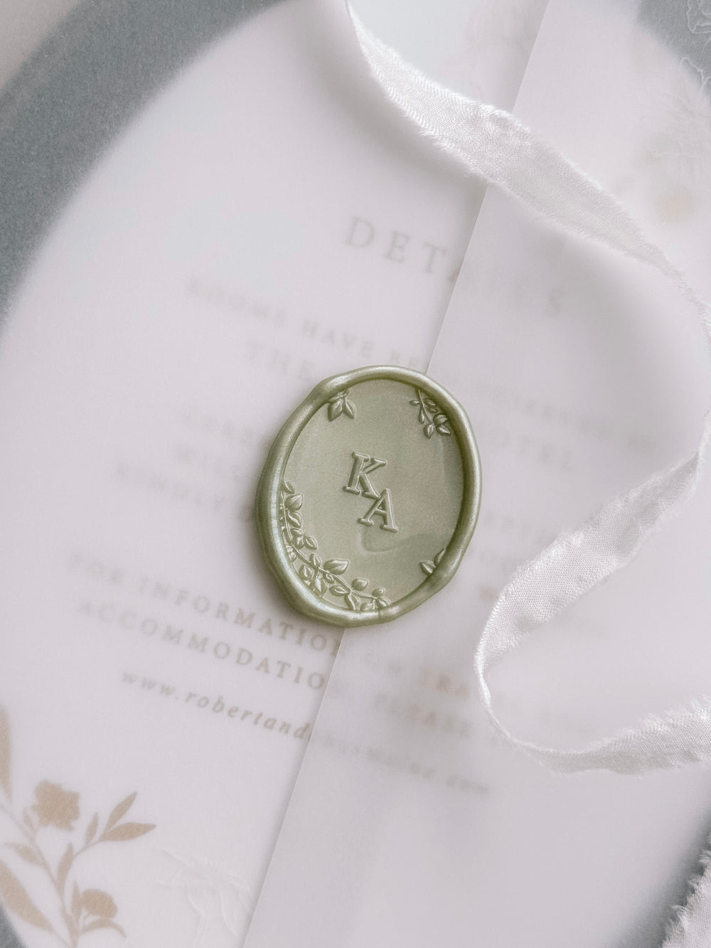 Light metallic green oval monogram wax seal personalized with initials framed by a botanical border with 3D engraving on a wedding invitation vellum jacket