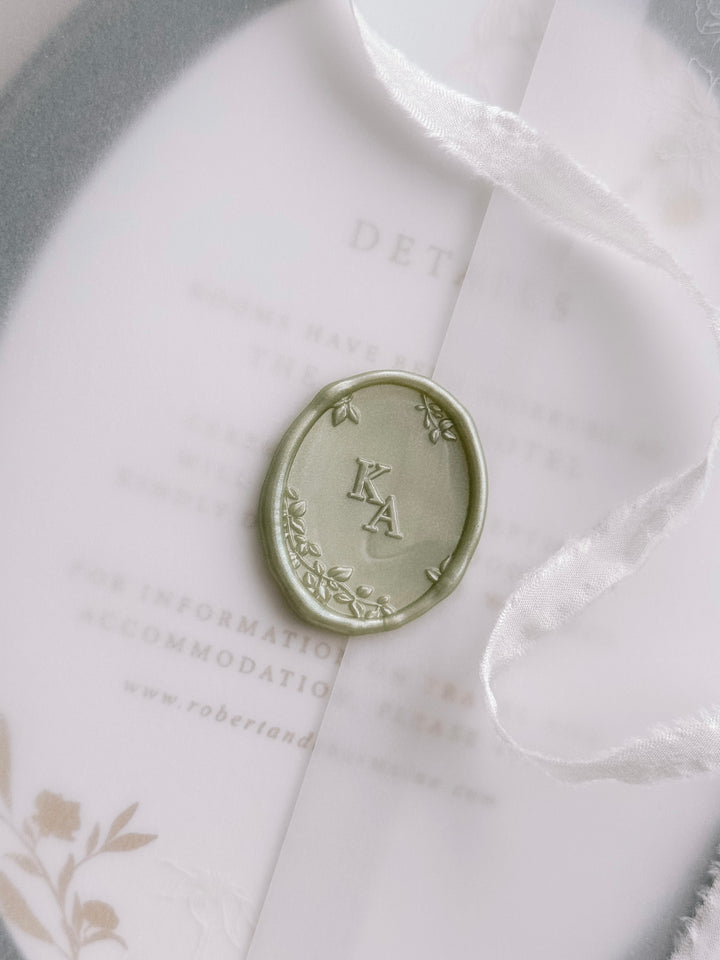 Light metallic green oval monogram wax seal personalized with initials framed by a botanical border with 3D engraving on a wedding invitation vellum jacket