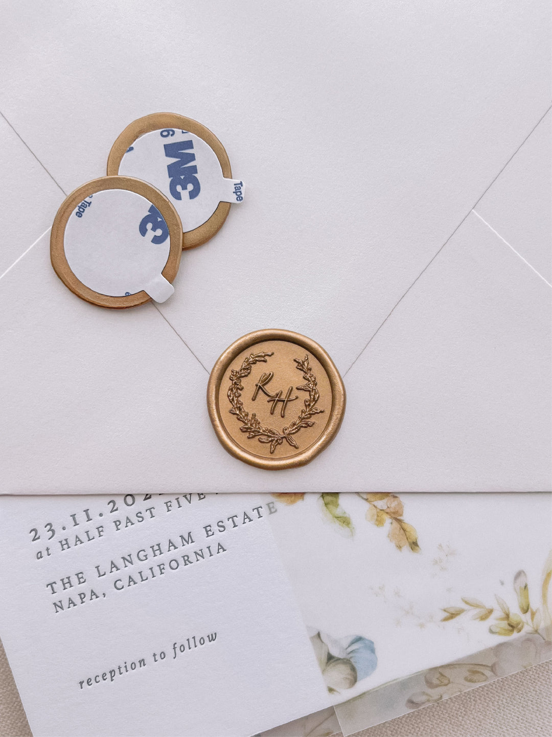 botanical wreath monogram gold wax seal stickers with self-adhesive backings on a wedding invitation, beige envelope, and floral vellum jacket