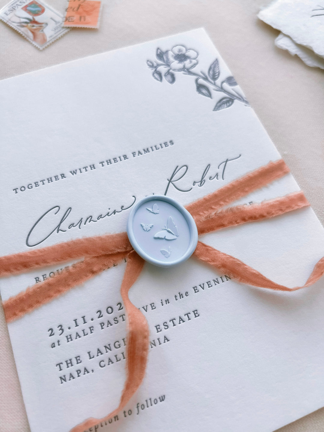 Light blue 3D oval butterflies wax seal on a white wedding invitation card tied with a narrow terracotta silk ribbon 