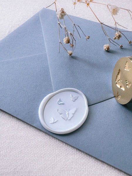 Pearl white oval wax seal featuring design of butterflies with 3D engraving details on a dusty blue envelope