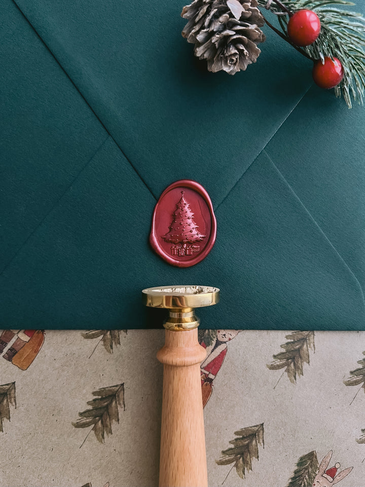 Dark metallic red oval Christmas tree wax seal with 3D engraved details on a dark green envelope with a wax stamp head and wooden handle on a festive background