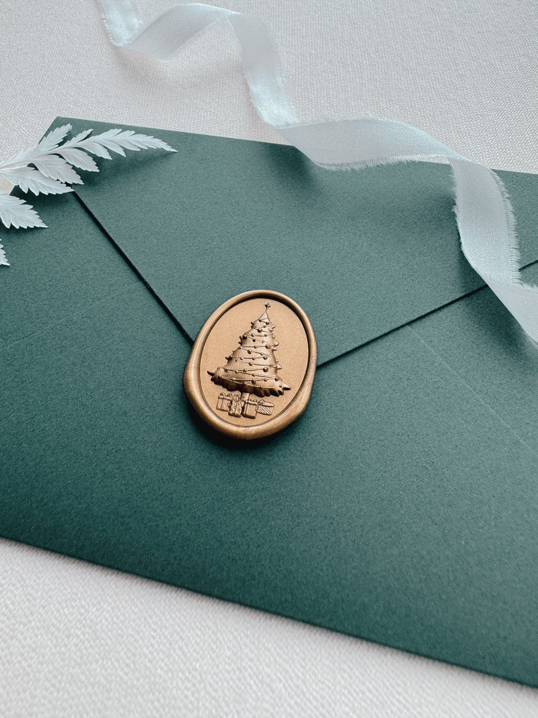 Gold Christmas tree oval wax seal with 3D engraved detail on a dark green envelope on a white background