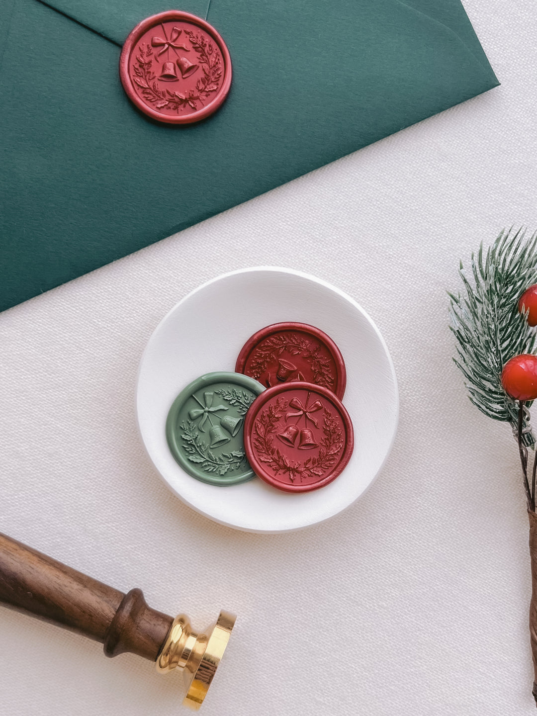 Sage green and dark metallic red 3D Christmas wreath and jingle bells wax seals on a dark green wedding envelope and a white ceramic dish next to a wax stamp head with dark wooden handle