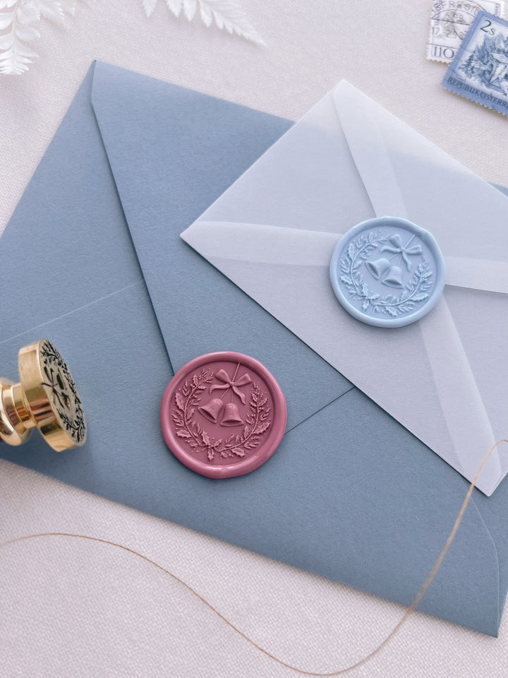 Light blue and rose pink 3D Christmas wreath and jingle bells wax seals on dusty blue envelope and mini vellum envelopes next to a wax stamp head