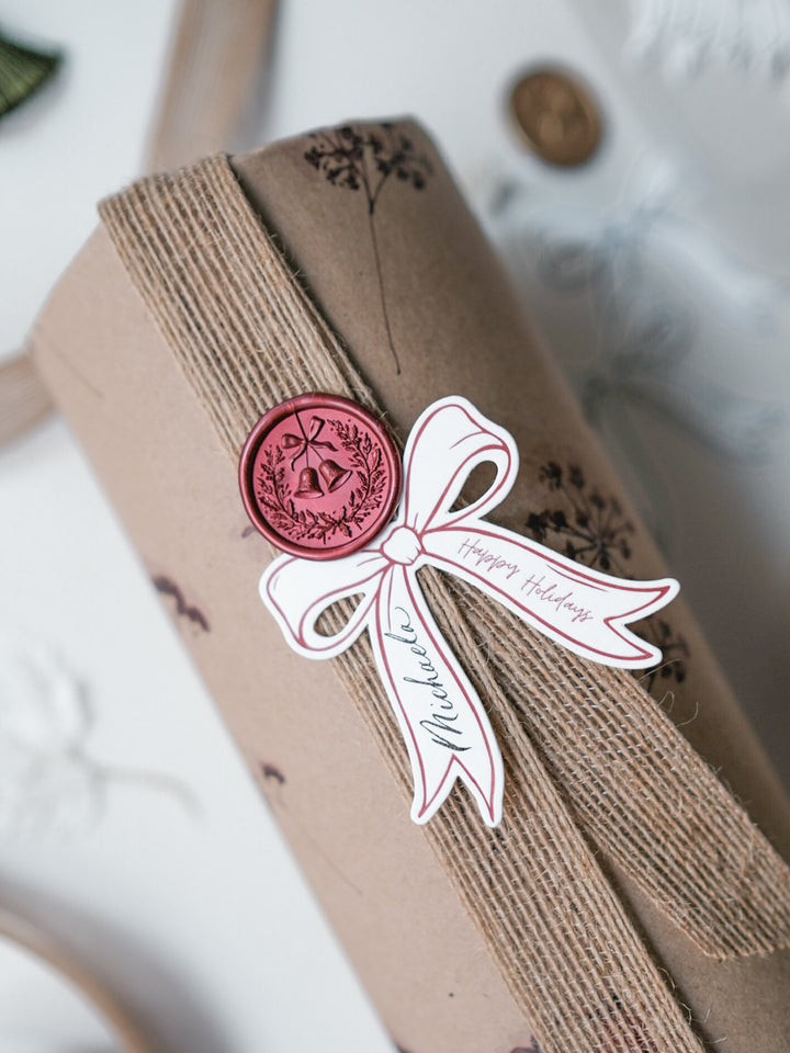 Dark metallic red Christmas wreath and jingle bells wax seal on a gift wrapped in brown kraft paper with botanical print and a ribbon bow die-cut personalized gift tag
