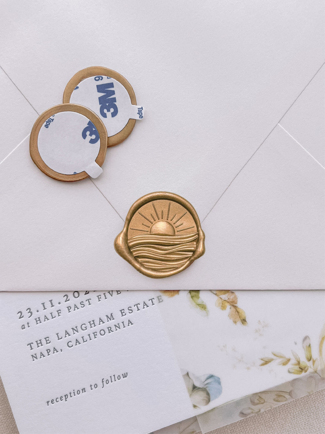 Gold 3D sun and ocean wax seal stickers with self-adhesive backings on a wedding invitation, beige envelope, and vellum jacket