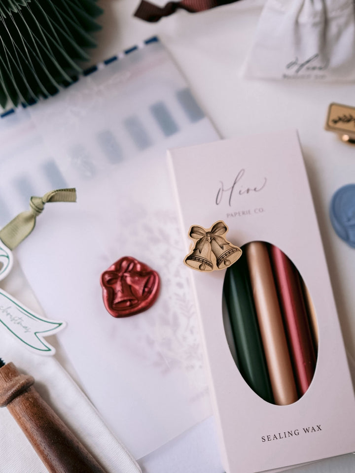 Dark metallic red die-cut jingle bells wax seal on a botanical vellum jacket, with the brass stamp head and a boxed set of sealing wax sticks in green, gold and red.