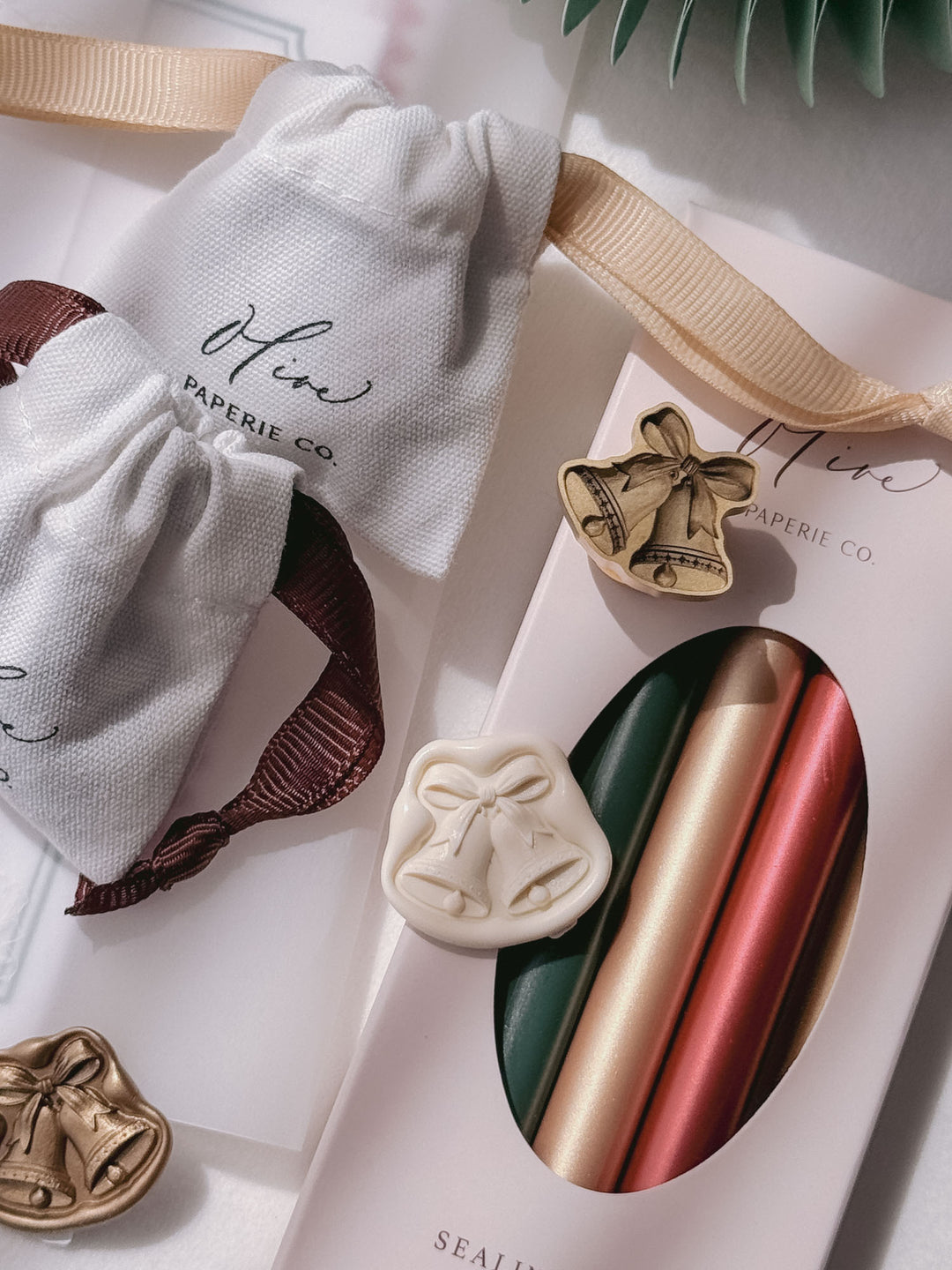 Off-white, gold and die-cut brass jingle bells wax seals arranged beside Olive Paperie Co. linen pouches and a boxed set of green, gold and red sealing wax sticks.