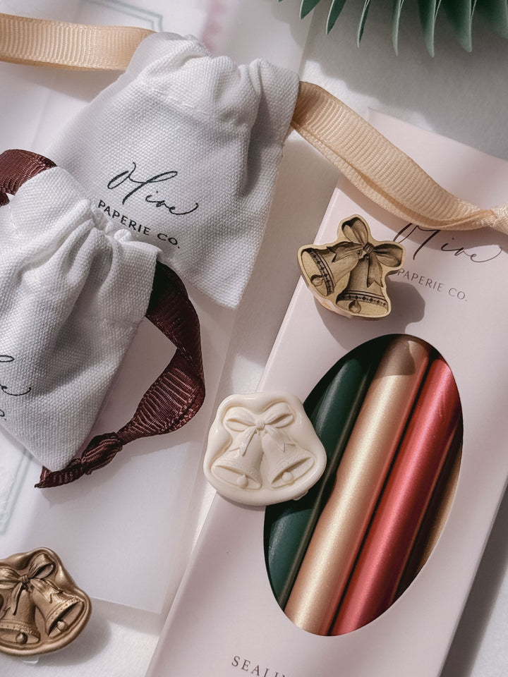 Off-white, gold and die-cut brass jingle bells wax seals arranged beside Olive Paperie Co. linen pouches and a boxed set of green, gold and red sealing wax sticks.