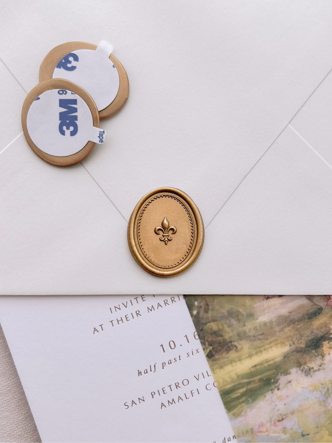 Gold 3D fleur de lis oval wax seal stickers with self-adhesive backings on a wedding invitation, beige envelope, and vellum jacket