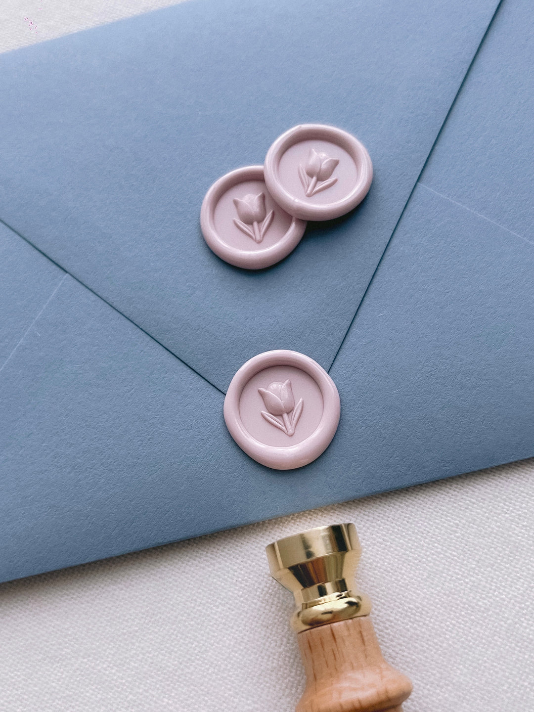 light pink 3D mini flower wax seals on a dusty blue wedding envelope next to a wax stamp head with light wood handle