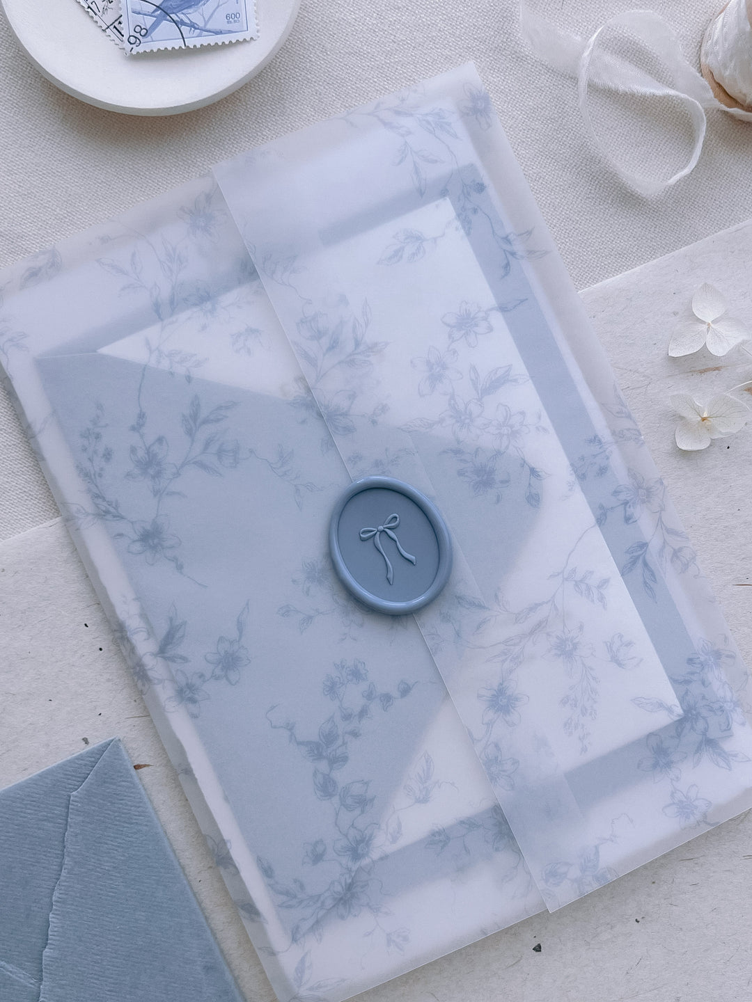 Dusty blue hand-drawn ribbon bow oval wax seal on a blue floral print wedding invitation vellum jacket 