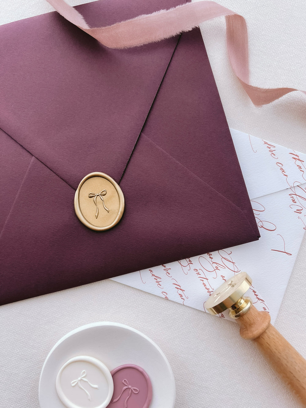 Gold 3D hand-drawn ribbon bow oval wax seal on a burgundy square envelope next to a wax stamp head with a wooden handle