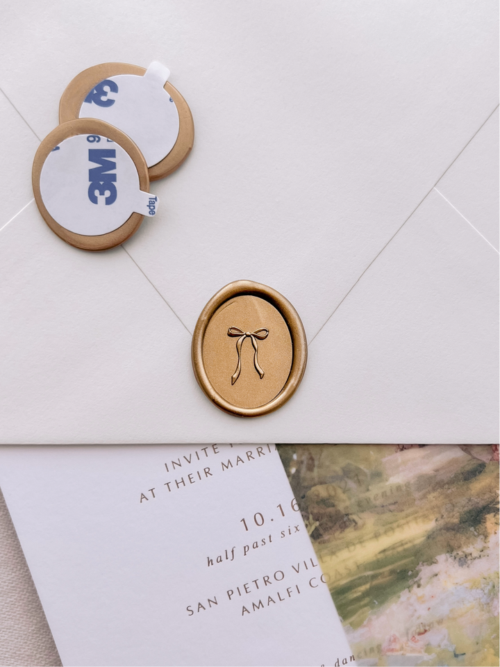 Gold 3D hand-drawn bow oval wax seal stickers with self-adhesive backings on a wedding invitation, beige envelope, and vellum jacket