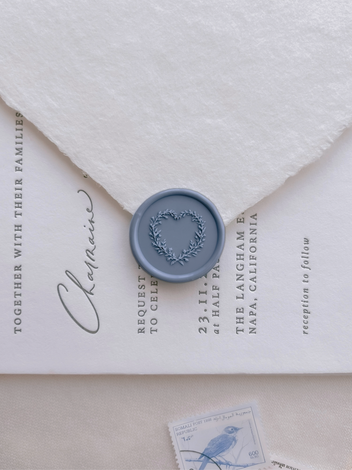Dusty blue heart shaped botanical wreath wax seal on a white envelope over a white wedding invitation card 