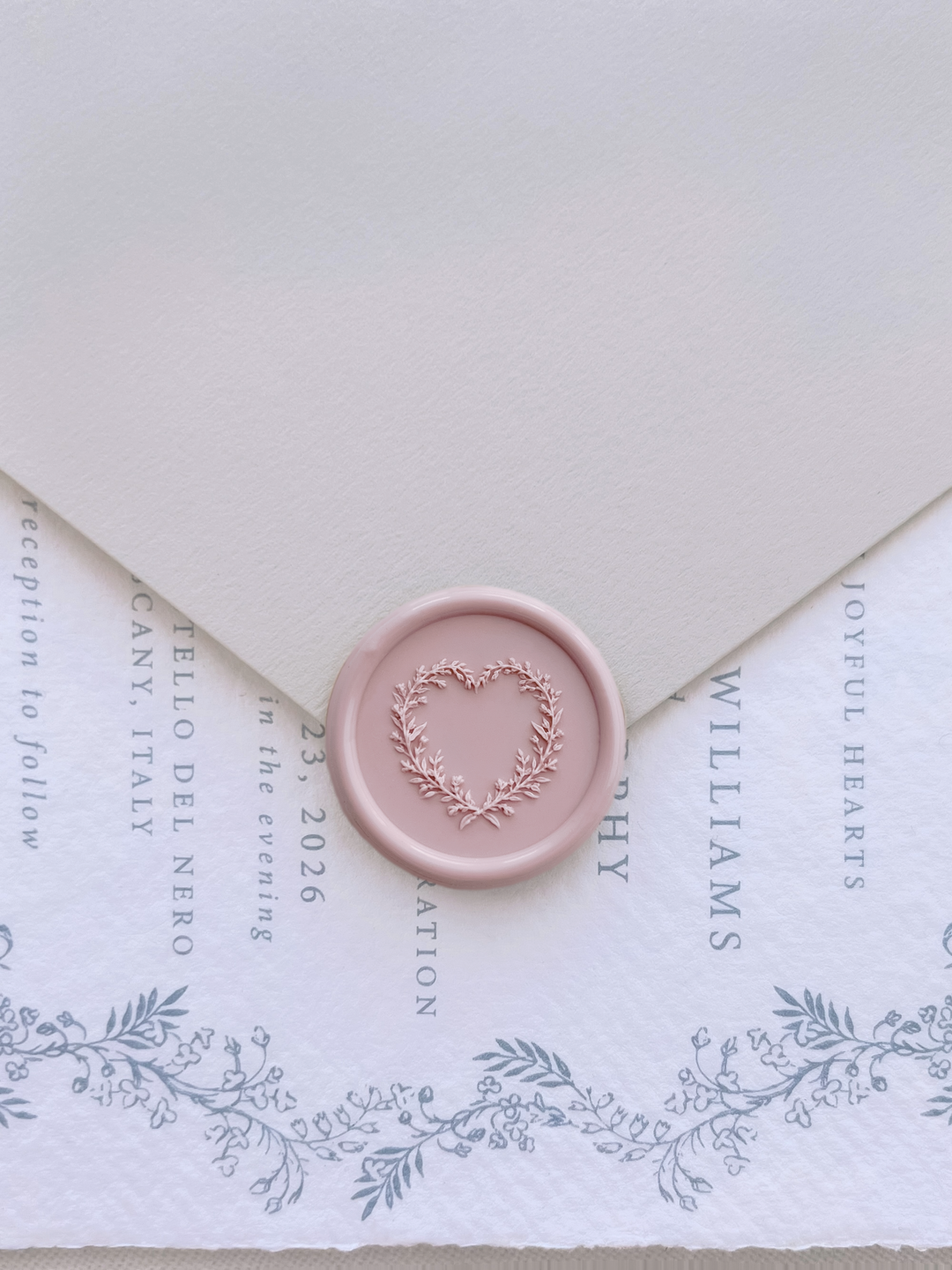 Light pink 3D heart shaped botanical wreath wax seal on a beige envelope over a dusty blue botanical wedding invitation card