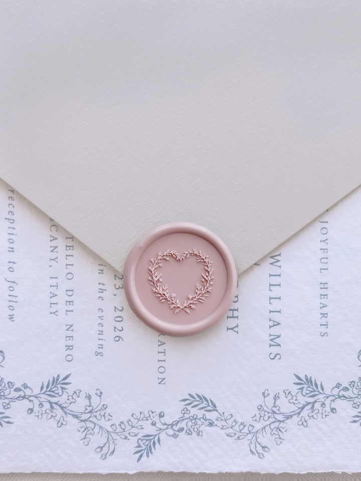 Light pink 3D heart shaped botanical wreath wax seal on a beige envelope over a dusty blue botanical wedding invitation card
