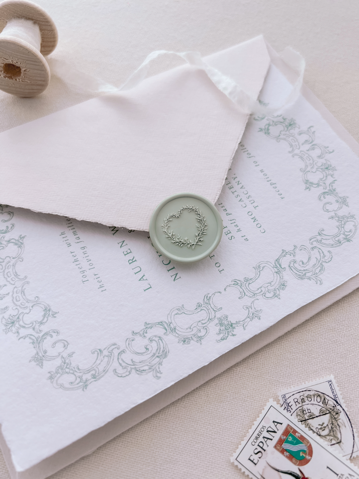 Light sage green heart shaped floral wreath wax seal on a pale pink envelope over a green vintage border wedding invitation card