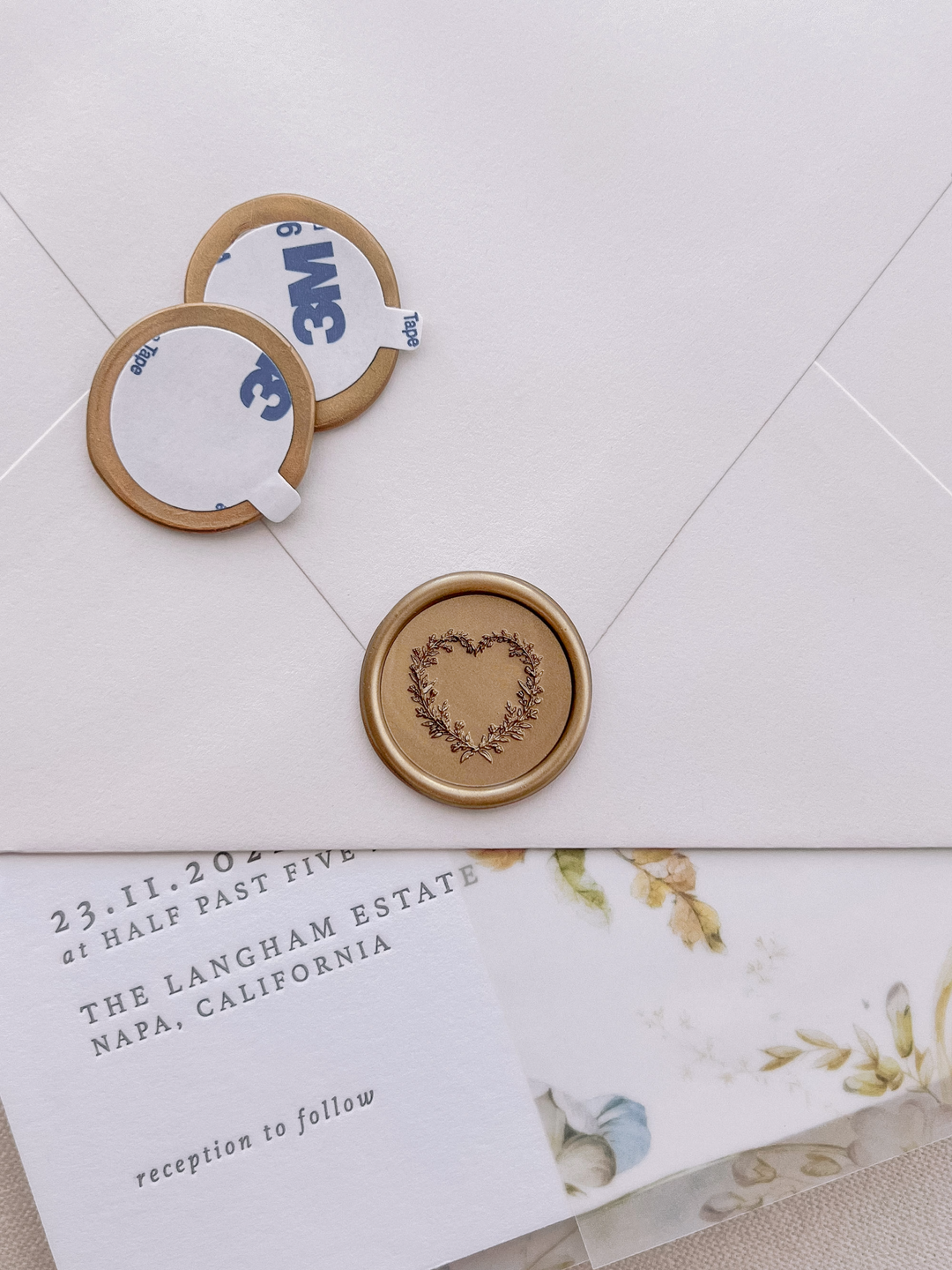 Gold heart-shaped botanical wreath wax seal stickers with self-adhesive backings on a beige envelope, floral vellum jacket and wedding invitation card 