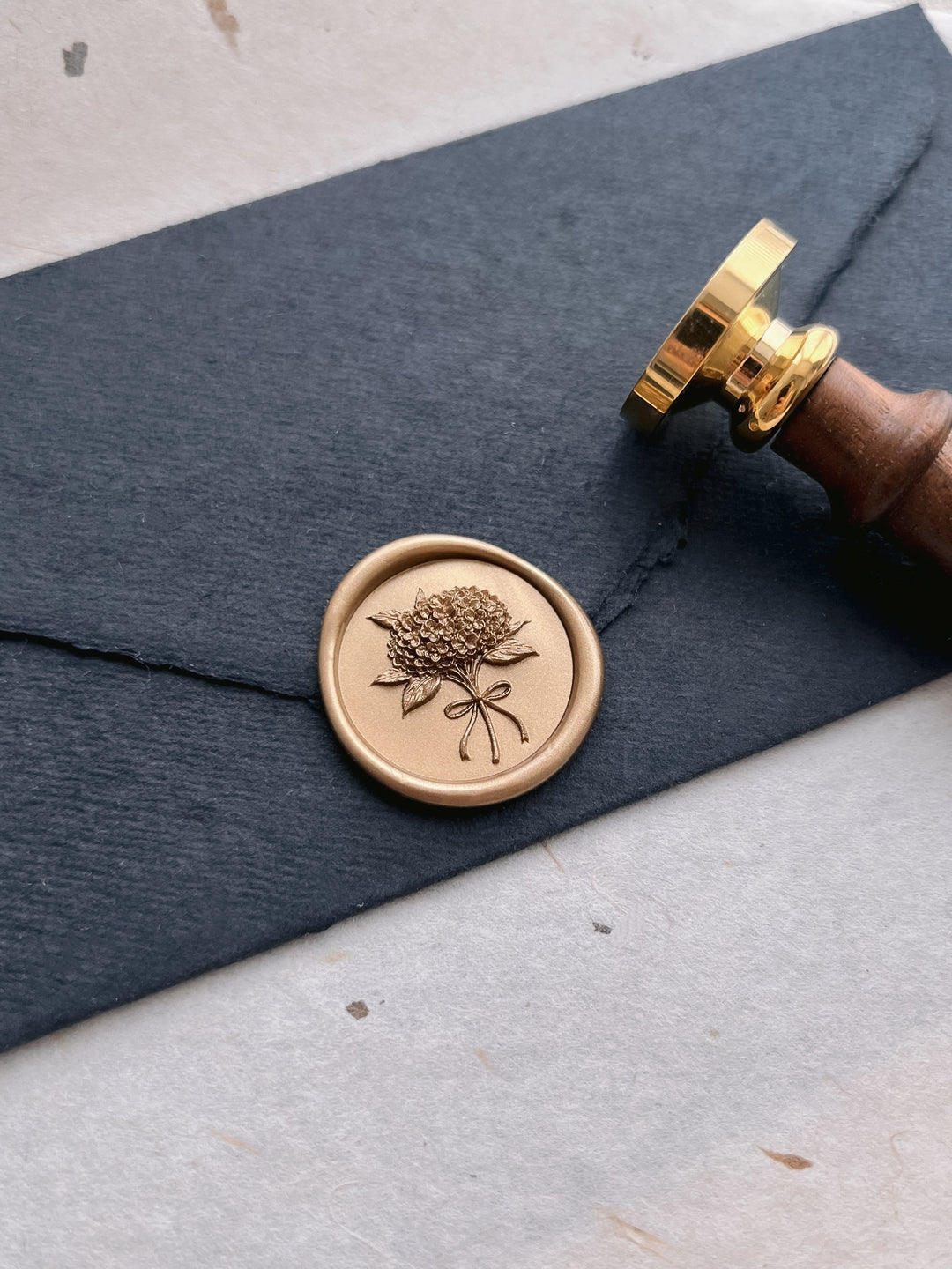 Golden bronze hydrangea flower bouquet wax seal on a black handmade paper envelope next to a brass wax stamp head and wooden handle 