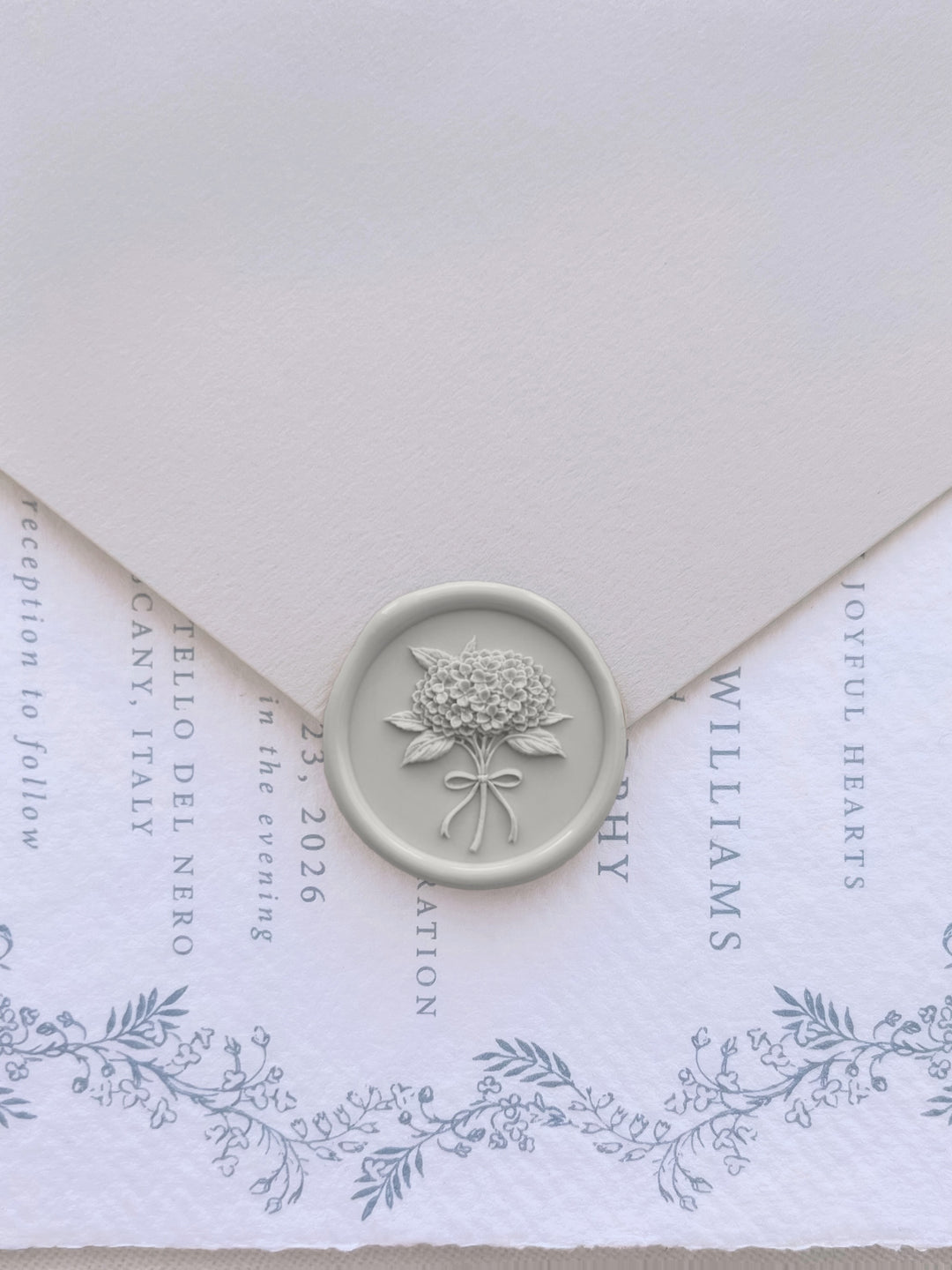 Grey hydrangea flower bouquet wax seal on a beige envelope over a blue botanical wedding invitation card 