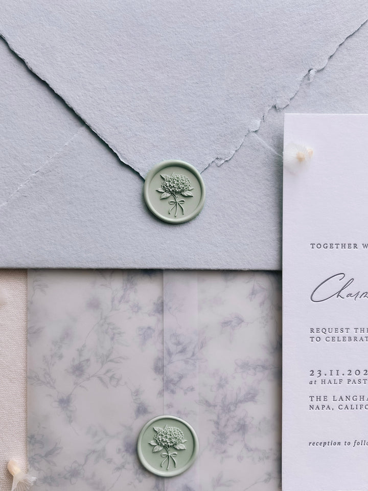 Light sage green 3D hydrangea bouquet wax seals on a light blue textured envelope and a gray floral vellum jacket next to a wedding invitation card