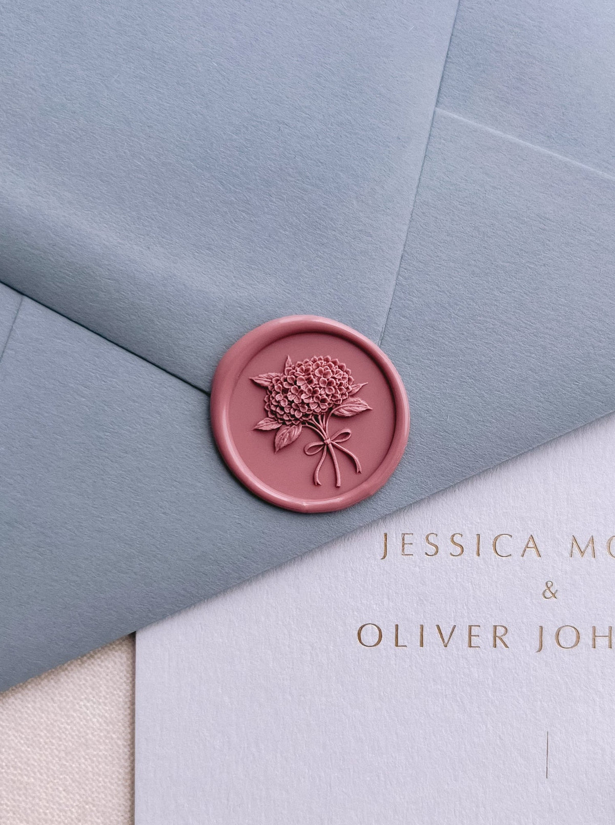 Rose colored 3D hydrangea bouquet wax seal on a dusty blue envelope and light blue wedding invitation card