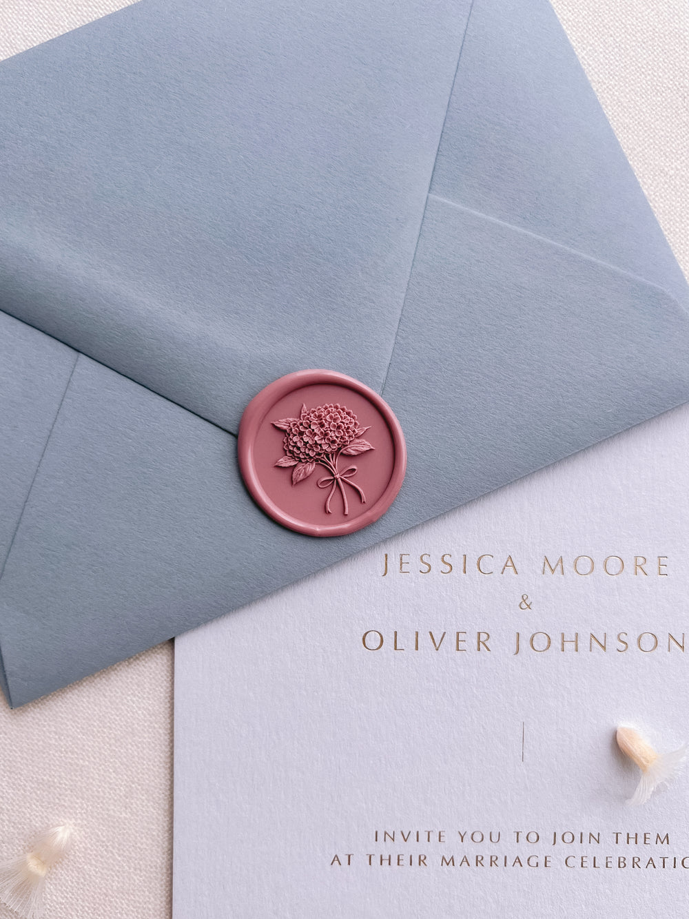 Rose pink hydrangea bouquet wax seal on a dusty blue envelope over a light blue gold foil wedding invitation card