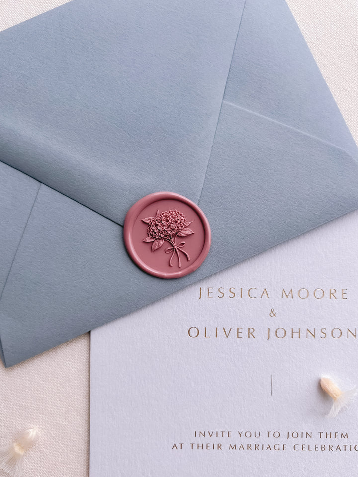 Rose pink hydrangea bouquet wax seal on a dusty blue envelope over a light blue gold foil wedding invitation card