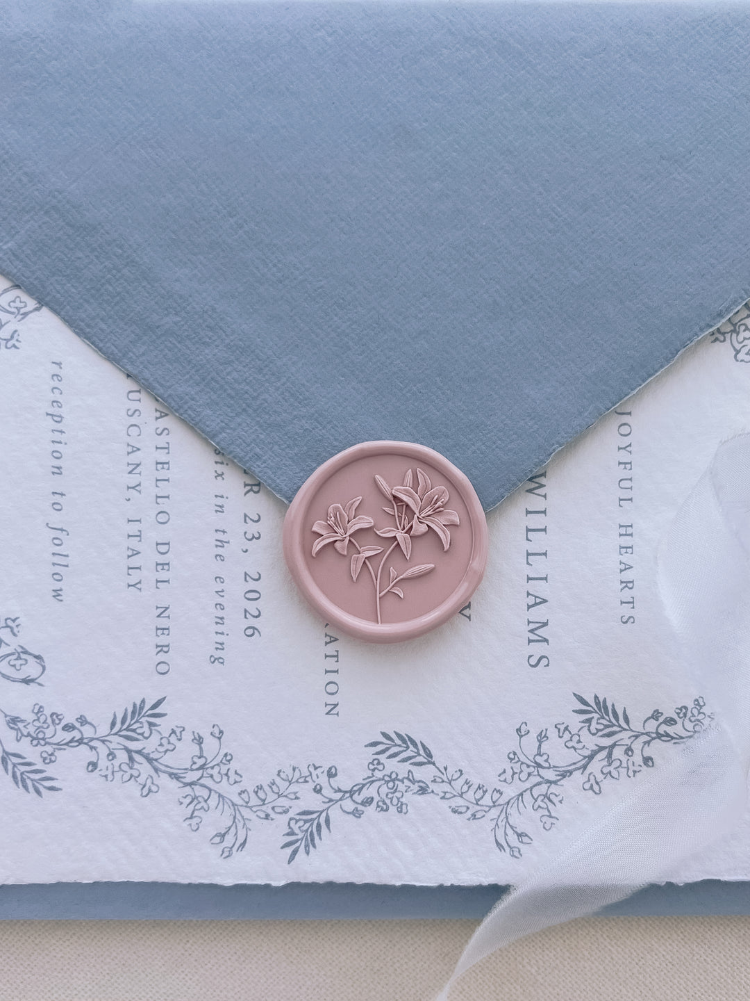 Light pink 3D lilies floral wax seal on a dusty blue envelope over a wedding invitation card