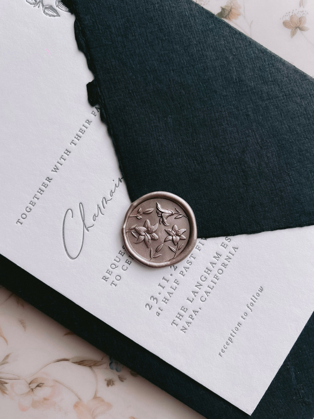 Light bronze 3D lily flowers wax seal on a black deckle-edged handmade paper envelope over a white wedding invitation card