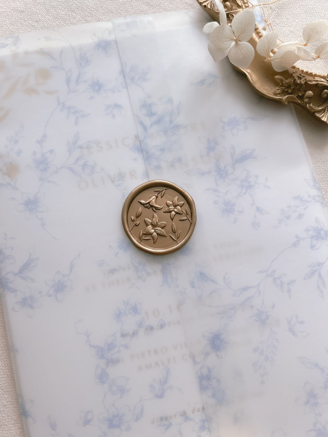 gold 3D lily flowers wax seal on a blue floral print vellum jacket over a wedding invitation card