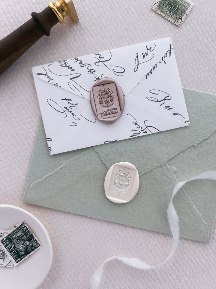 light bronze and white festive holiday envelope seals featuring design of the phrase Merry Christmas and an ornament design on sage green and white envelopes