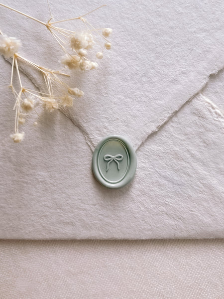 Light sage green hand-drawn ribbon bow mini oval wax seal on a white textured envelope
