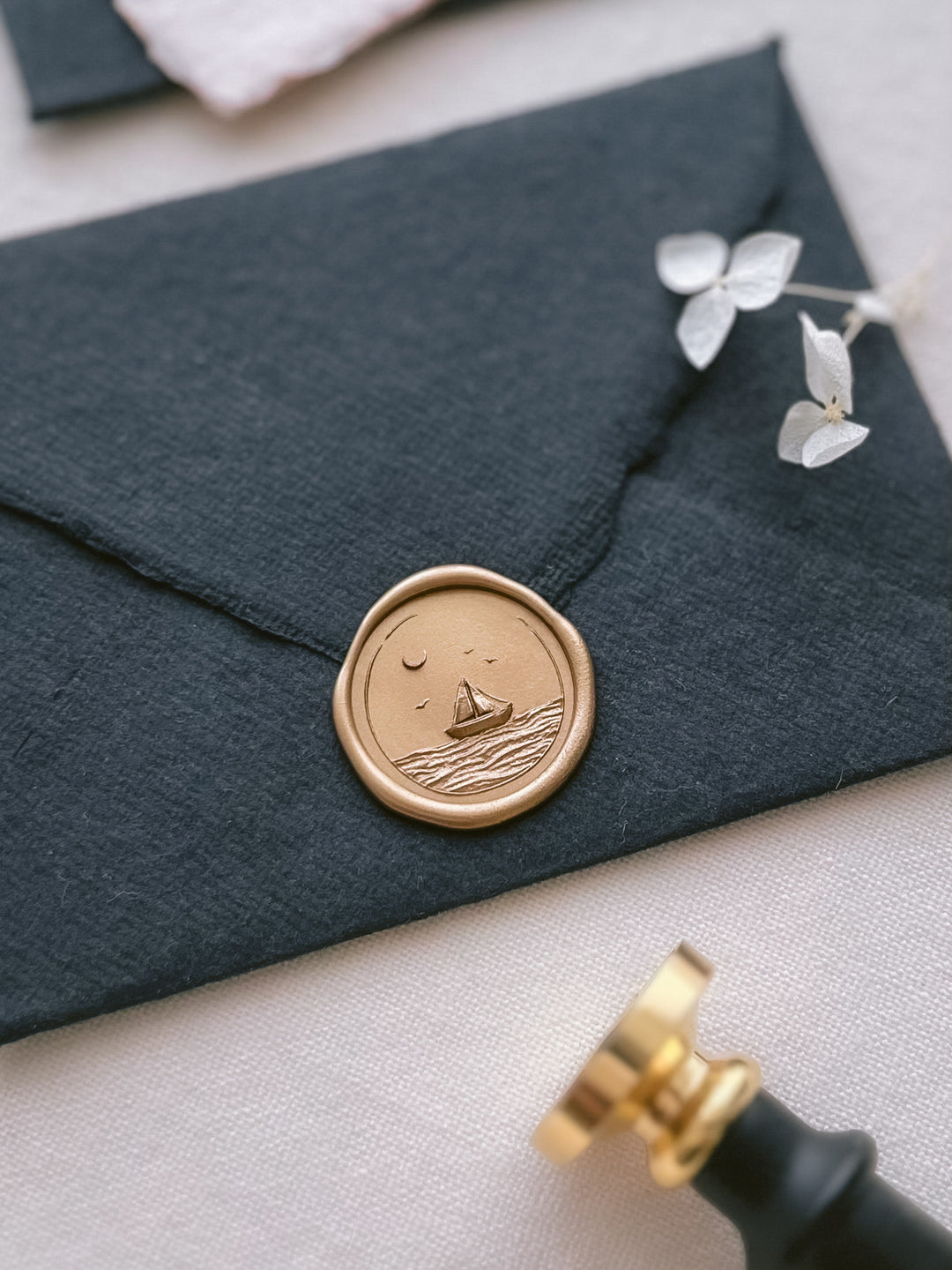 Gold 3D engraved sailboat and ocean waves wax seal on a black handmade paper envelope next to a brass wax stamp head and black handle 