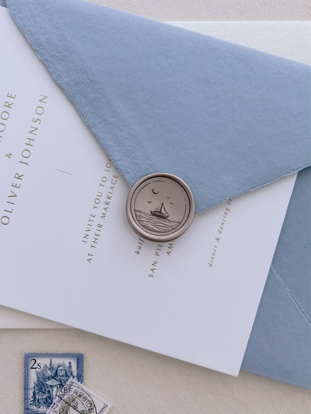Light bronze 3D-engraved sailboat coastal wax seal on a dusty blue handmade paper envelope over a wedding invitation card