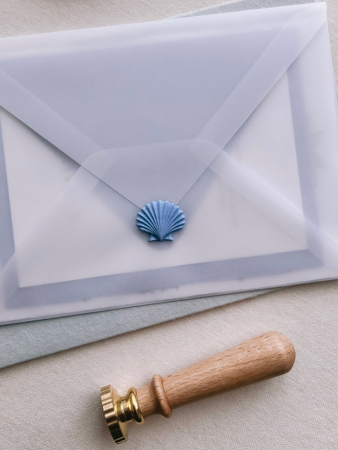 French blue 3D seashell wax seal on a wedding invitation vellum envelope placed next to a wax seal stamp with wooden handle
